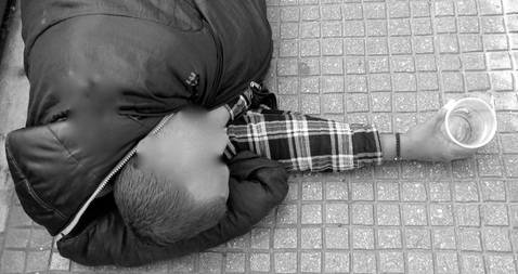 Asunnoton makaa maassa makuupussissa. Hän on ojentanut käden, jossa on muovimuki. Jan Holmberg Mainio blogi janholmberg.weebly.com Copyright Jan Holmberg 